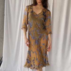 May include: A sheer, knee-length dress in a mustard yellow hue, adorned with a floral pattern in shades of purple and blue. The dress features a V-neckline, ruffled sleeves, and a ruffled hem. The model is wearing gold-colored heels.