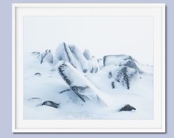 May include: A snowy landscape with large, dark rocks partially covered in white snow. The rocks are clustered together in a natural formation. The background is a hazy white, suggesting a foggy or snowy day.