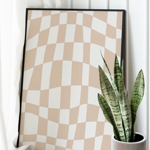 May include: A framed print with a wavy checkered pattern in beige and white. The print is leaning against a white wall next to a potted snake plant in a white planter.