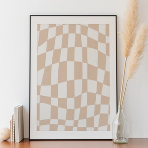 May include: A black framed print featuring a beige and white checkered pattern. The print is hanging on a white wall above a wooden dresser with two drawers. A vase containing dried pampas grass is on the dresser.