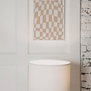 May include: A white lampshade with a wooden tripod base sits on a white surface in front of a framed print with a brown and white checkered pattern.