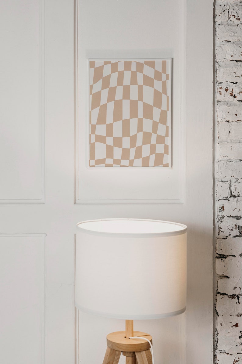 May include: A white lampshade with a wooden tripod base sits on a white surface in front of a framed print with a brown and white checkered pattern.