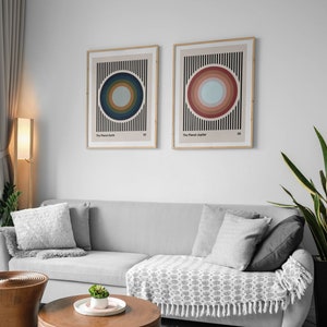 May include: Two framed abstract art prints with colorful concentric circles on a striped background. The prints are titled "The Planet Earth" and "The Planet Jupiter". The prints are hanging on a white wall above a gray sofa with pillows and a throw blanket.