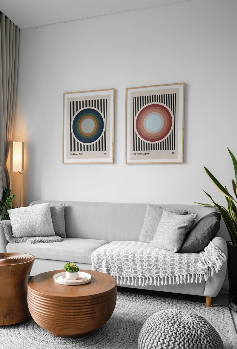May include: Two framed abstract art prints with colorful concentric circles on a striped background. The prints are titled "The Planet Earth" and "The Planet Jupiter". The prints are hanging on a white wall above a gray sofa with pillows and a throw blanket.