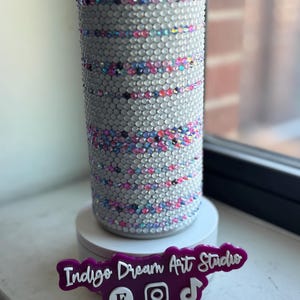 May include: A cylindrical tumbler fully covered in clear, reflective rhinestones with horizontal bands of pink, blue, and purple crystals. A purple sign in front reads Indigo Dream Art Studio with icons for Etsy, Instagram, and TikTok.