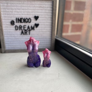 Può includere: Due sculture di torso in resina. La scultura più grande è viola con una parte superiore rosa e glitterata. La scultura più piccola è rosa con glitter. Le sculture sono su una superficie bianca. Una lavagna a lettere incorniciata sullo sfondo recita "@ INDIGO DREAM ART".