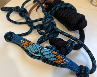 Può includere: Un capezzone per cavalli in corda blu e nera con una fascetta nasale decorativa con perline con un motivo a piume turchesi e arancioni. Il capezzone include un cinturino in pelle e una sezione di corda nera.
