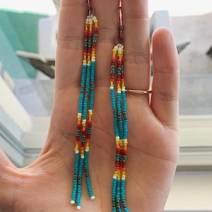 May include: Handmade beaded earrings with a copper hook. The earrings feature turquoise, red, yellow, and white beads. The beaded strands hang down, creating a fringe effect. The earrings are held in a hand.