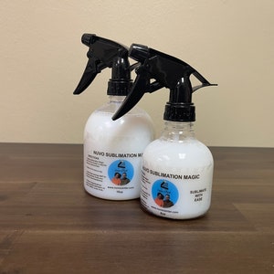 Puede incluir: Dos botellas de spray transparentes llenas de un líquido blanco, etiquetadas como "NUVO Sublimation Magic". Cada botella tiene una boquilla de pulverización negra. Las etiquetas incluyen el texto "Sublimate with Ease" y una dirección web.