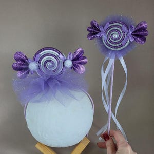 Pode incluir: Dois acessórios roxos com tema de doces. Um é uma tiara com um grande design de doce, tule e uma base de bola branca. O outro é uma varinha com um design de doce semelhante, glitter e fitas. Os designs de doces apresentam um centro em espiral.