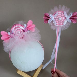 Pode incluir: Um conjunto de faixa de cabelo e varinha mágica com tema de doces rosa e branco. A faixa de cabelo apresenta um grande design de doce em espiral com detalhes rosa e branco, rodeado por tule e enfeites rosa. A varinha tem um design de doce semelhante e uma pega rosa.