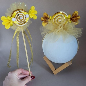 Pode incluir: Duas decorações extravagantes com tema de doces. Uma é uma varinha com uma alça de fita dourada e um design de doce amarelo e dourado. A outra é uma esfera branca com um topo de doce, colocada em um suporte de madeira. Ambas têm detalhes em amarelo.