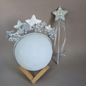 Puede incluir: Una diadema de estrella plateada con una varita de brillo plateada. La diadema está decorada con múltiples estrellas plateadas y la varita tiene una estrella plateada en la parte superior con una cinta blanca.