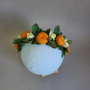 Könnte beinhalten: Eine Filzkrone mit Orangenblüten, grünen Blättern und weißen Blüten. Die Krone befindet sich auf einer hellblauen Styroporkugel.