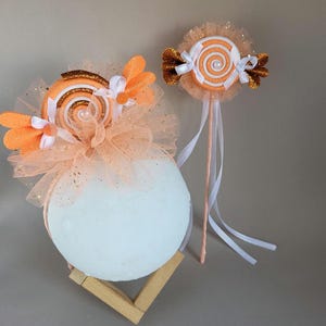 Pode incluir: Duas decorações extravagantes com tema de doces. Uma é uma tiara com um grande pirulito de redemoinho laranja e branco, adornado com detalhes laranja e tule. A outra é uma varinha com um design de pirulito semelhante, com fitas brancas e detalhes dourados.
