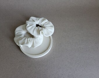May include: Two white fabric scrunchies on a white round ceramic dish.