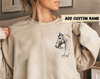May include: A beige sweatshirt with a black and white illustration of a horse's head on the front. The text "ADD CUSTOM NAME" is at the top of the image and the name "Axel" is below the horse's head.