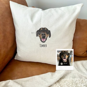 May include: A white pillow with a black and brown dog's face embroidered on it. The dog's name, "TIMBER", is embroidered below the face. A photo of the dog is on a white card in the bottom right corner of the image.