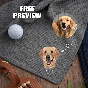 K&ouml;nnte beinhalten: Ein graues Golftuch mit einem gestickten Bild eines Golden Retriever-Hundes und dem Namen "KODA". Ein Golfball und ein Teehalter sind ebenfalls sichtbar. Der Text "FREE PREVIEW" ist oben links zu sehen.