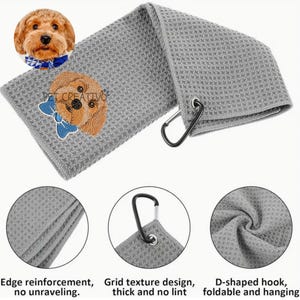K&ouml;nnte beinhalten: Graues Golfhandtuch mit Waffelstruktur und einem gestickten Hund mit blauer Schleife. Mit D-f&ouml;rmigem Haken zum Aufh&auml;ngen und dem Text "PET CREATIVO". Das Handtuch hat eine Kantenverst&auml;rkung und ein Gittermuster.