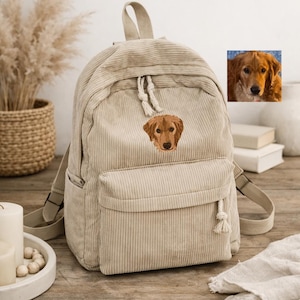 May include: Beige corduroy backpack with a front pocket and a small embroidered image of a golden retriever dog. The backpack has adjustable straps and a top handle. The background includes a wooden table and decorative items.