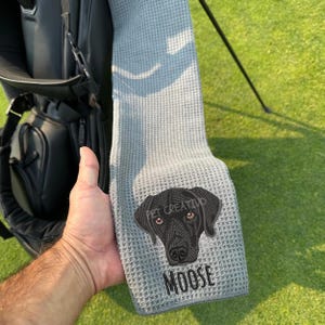 K&ouml;nnte beinhalten: Graues Golfhandtuch mit Waffelstruktur. Es zeigt ein gesticktes schwarzes Labrador Retriever-Gesicht mit dem Namen "MOOSE" darunter. Der Text "PET CREATIVO" befindet sich &uuml;ber dem Kopf des Hundes. Das Handtuch wird in der N&auml;he einer schwarzen Golftasche gehalten.