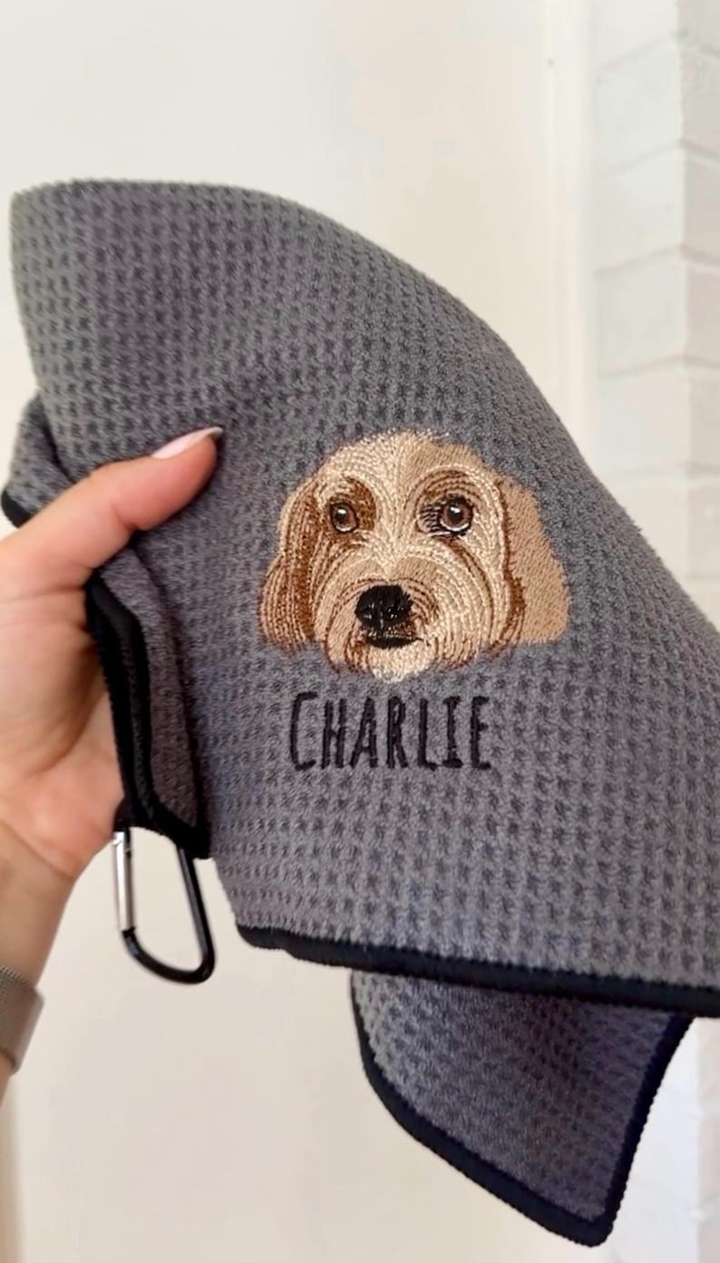 K&ouml;nnte beinhalten: Graues Waffelhandtuch mit schwarzem Rand und Metallclip. Das Handtuch zeigt ein gesticktes Hunde-Gesicht in Braun- und Beiget&ouml;nen, darunter der Name "CHARLIE" in Schwarz gestickt.
