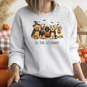 May include: A light gray crewneck sweatshirt featuring a graphic of five dogs in Halloween costumes, including a witch, a pumpkin, and a ghost. The text below the dogs reads, "SIT, STAY, GET SPOOKY!" The sweatshirt has long sleeves and a ribbed collar.