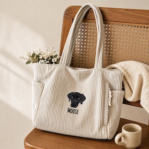 May include: A cream-colored corduroy tote bag featuring a vertical ribbed texture and two shoulder straps. The front displays an embroidered black dog face above the text MOOSE in black capital letters. The bag includes side pockets.