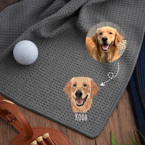 K&ouml;nnte beinhalten: Graues Golfhandtuch mit Waffelstruktur. Das Handtuch zeigt ein gesticktes Bild eines Golden Retriever-Hundes mit dem Namen "KODA" darunter. Ein Golfball liegt auf dem Handtuch. Ein rundes Foto eines Golden Retrievers ist ebenfalls vorhanden.