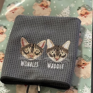 K&ouml;nnte beinhalten: Graues Waffelhandtuch mit gestickten Katzenk&ouml;pfen und den Namen "Wobbles" und "Waddle". Das Handtuch hat einen schwarzen Rand und einen Metallclip. Der Hintergrund zeigt Geschenkpapier mit Weihnachtsmann und Weihnachtsb&auml;umen.