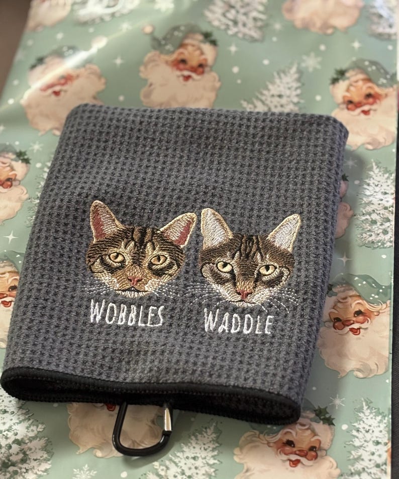 K&ouml;nnte beinhalten: Graues Waffelhandtuch mit gestickten Katzenk&ouml;pfen und den Namen "Wobbles" und "Waddle". Das Handtuch hat einen schwarzen Rand und einen Metallclip. Der Hintergrund zeigt Geschenkpapier mit Weihnachtsmann und Weihnachtsb&auml;umen.