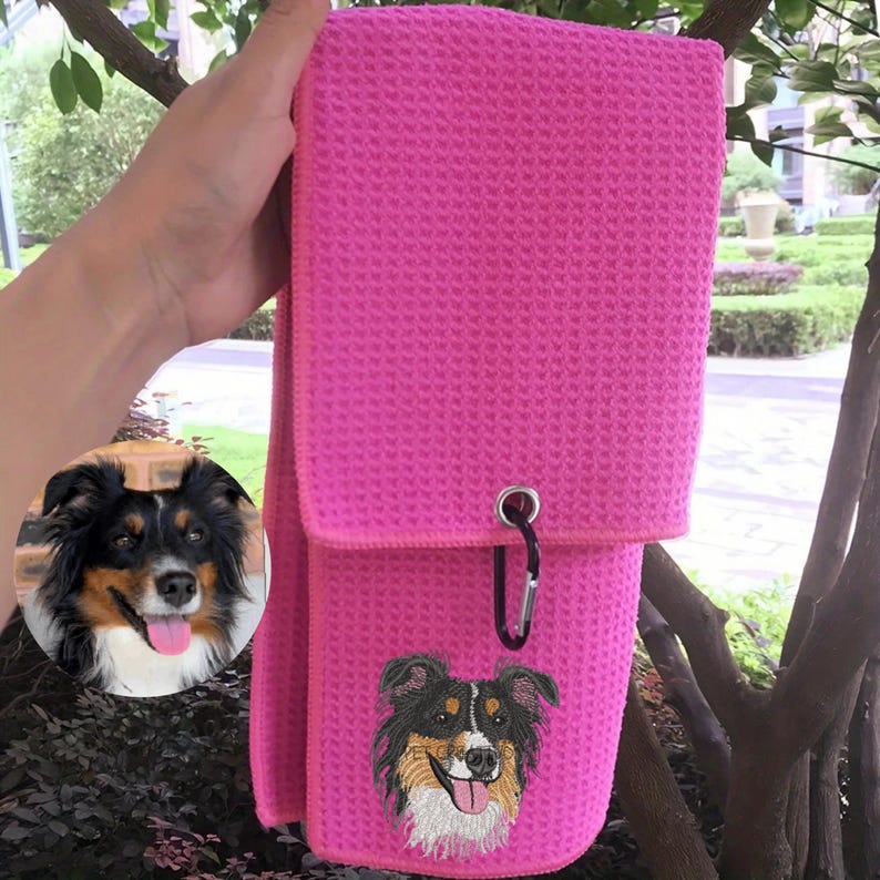 K&ouml;nnte beinhalten: Ein leuchtend pinkfarbenes Waffelhandtuch mit einem gestickten Hundebild. Der Hund hat schwarzes, braunes und wei&szlig;es Fell, mit herausgestreckter Zunge. Eine silberne &Ouml;se und ein schwarzer Karabinerhaken sind am Handtuch befestigt.