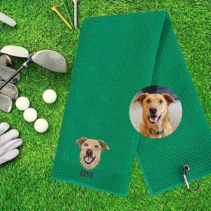 K&ouml;nnte beinhalten: Gr&uuml;nes Golfhandtuch mit einem gestickten Hundegesicht und dem Namen "HANK" darunter. Ein kreisf&ouml;rmiges Bild eines Hundes befindet sich auf dem Handtuch. Golfschl&auml;ger, Golfb&auml;lle und Tees im Hintergrund.