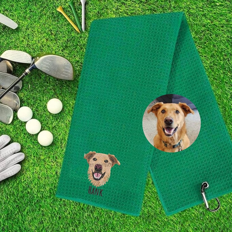 K&ouml;nnte beinhalten: Gr&uuml;nes Golfhandtuch mit einem gestickten Hundegesicht und dem Namen "HANK" darunter. Ein kreisf&ouml;rmiges Bild eines Hundes befindet sich auf dem Handtuch. Golfschl&auml;ger, Golfb&auml;lle und Tees im Hintergrund.