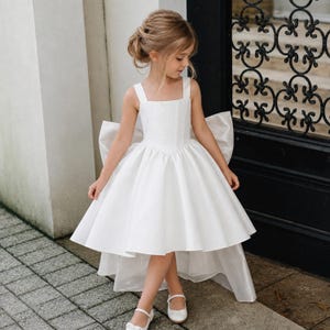 Könnte beinhalten: Ein weißes ärmelloses Kleid mit quadratischem Ausschnitt, eng anliegendem Oberteil und einem voluminösen Rock mit asymmetrischem Saum. Die Rückseite des Kleides ist mit einer großen, auffälligen Schleife verziert. Der Stoff ist glatt und einfarbig weiß.