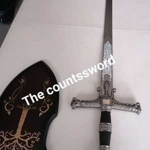 May include: A decorative sword with a silver blade and ornate hilt, featuring a black handle and a gold emblem. The sword is displayed with a black wooden plaque with a tree design. The text "the countssword" is visible.