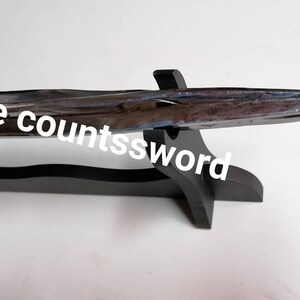 May include: A polished metal sword with a sharp point, resting on a black wooden stand. The sword has a dark handle with a gold-colored band near the blade. The text "The countsssword" is written on the image.