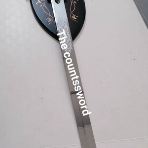 May include: A decorative sword with a silver blade and ornate hilt, mounted on a black shield-shaped plaque. The blade has the text "The countssword". The hilt and plaque feature gold-colored accents.