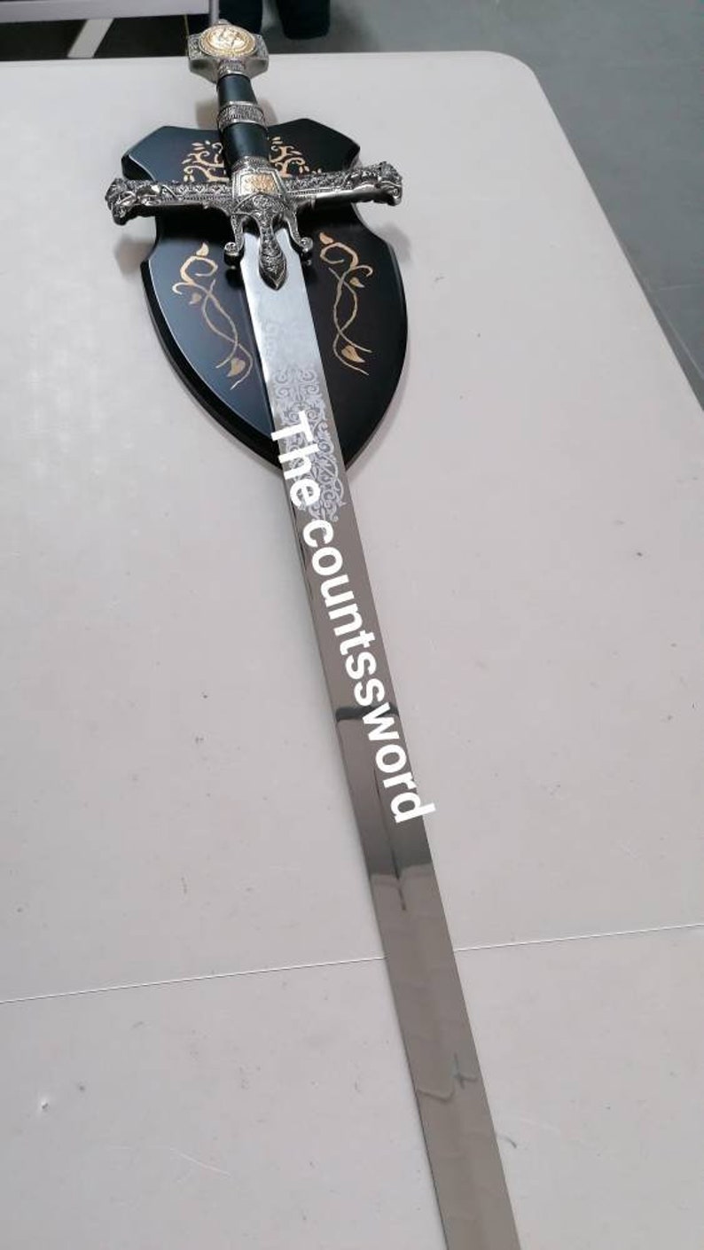 May include: A decorative sword with a silver blade and ornate hilt, mounted on a black shield-shaped plaque. The blade has the text "The countssword". The hilt and plaque feature gold-colored accents.