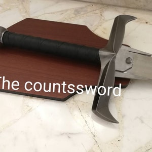 May include: A medieval-style sword with a silver blade and a black-wrapped handle. The sword is mounted on a dark brown wooden plaque. The text "The countssword" is visible on the plaque.