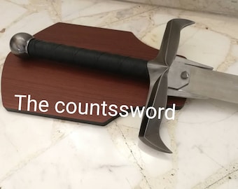 Puede incluir: Una espada de estilo medieval con una hoja plateada y un mango envuelto en negro. La espada está montada sobre una placa de madera marrón oscuro. El texto "The countssword" es visible.