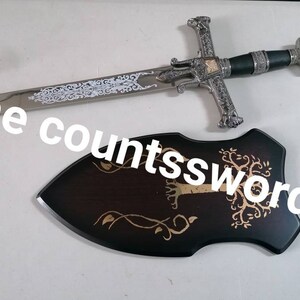 May include: A decorative sword with a silver blade and ornate hilt, accompanied by a wooden plaque. The sword features intricate designs and a black handle. The plaque is dark brown with a gold tree and vine design. The text "The countssword" is visible.