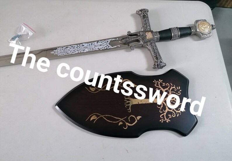 May include: A decorative sword with a silver blade and ornate hilt, accompanied by a wooden plaque. The sword features intricate designs and a black handle. The plaque is dark brown with a gold tree and vine design. The text "The countssword" is visible.