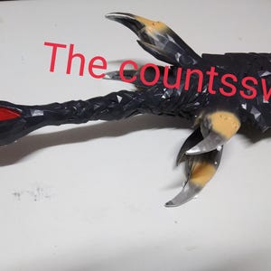 Może przedstawiać: Czarny i srebrny miecz o kształcie pazura. Miecz ma czerwony akcent w pobliżu czubka i napis "The countssword" w kolorze czerwonym. Ostrze ma teksturowany wygląd.