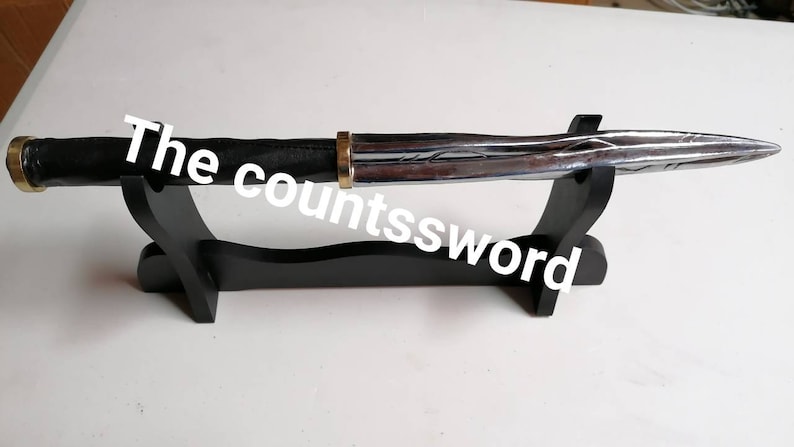 May include: A silver and black dagger with a black handle and a gold-colored ring. The dagger is displayed on a black wooden stand. The text "The countssword" is written across the image.