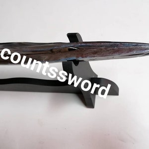 May include: A polished metal sword with a black handle and a gold-colored ring, resting on a black wooden stand. The text "The countssword" is written on the image.