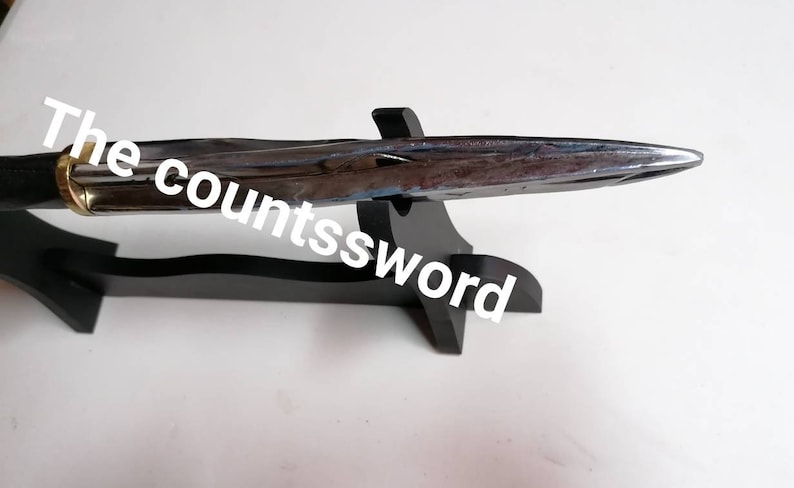 May include: A polished metal sword with a black handle and a gold-colored ring, resting on a black wooden stand. The text "The countssword" is written on the image.