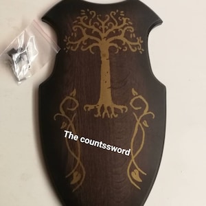 Könnte beinhalten: Dunkelbraune Holzschwertplakette mit goldenem Baumdesign und Rankenverzierungen. Der Text "The countssword" ist auf der Plakette zu sehen. Enthält einen kleinen Beutel mit Hardware.