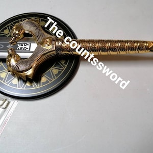 May include: A decorative sword with a gold-colored handle and a silver blade. The handle features intricate detailing and a dragon-like design. The sword rests on a black and gold circular stand. The text "The countssword" is visible.